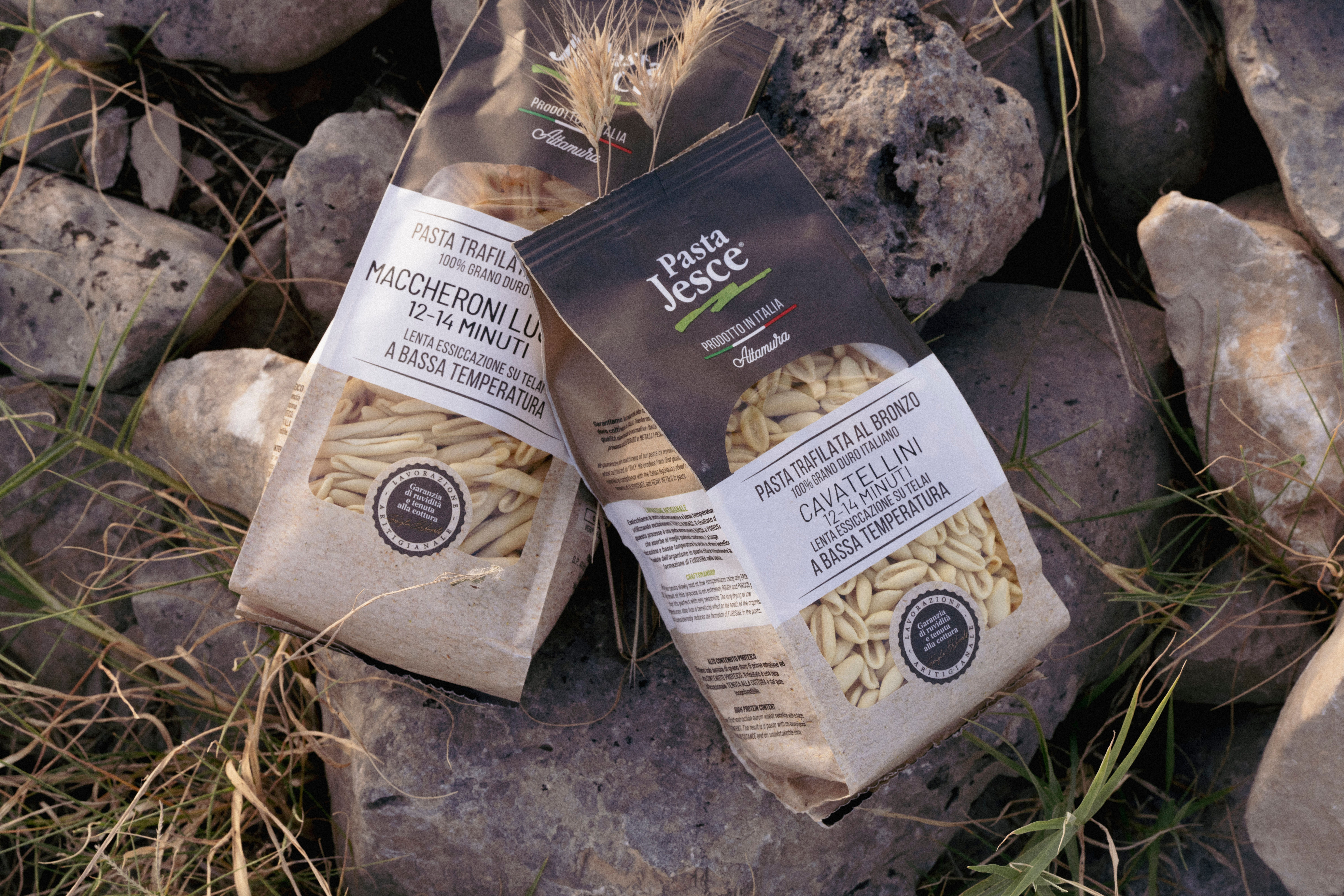 Zwei Packungen Pasta Jesce (Maccheroni, Cavatellini) aus Altamura auf Felsen und trockenem Gras mit Weizenähren.
