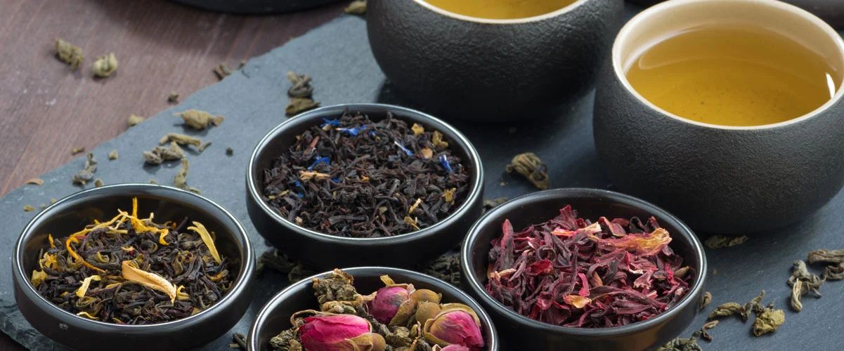 Vier Schalen mit losem Tee: Schwarztee mit gelben Blüten, Schwarztee mit blauen Blüten, Grüntee mit Rosenknospen,…