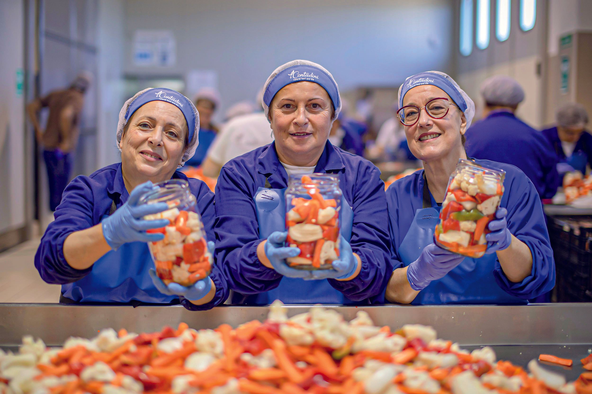 Drei lächelnde Frauen von Contadini in blauen Uniformen halten Gläser mit geschnittenem Gemüse in der Produktion.