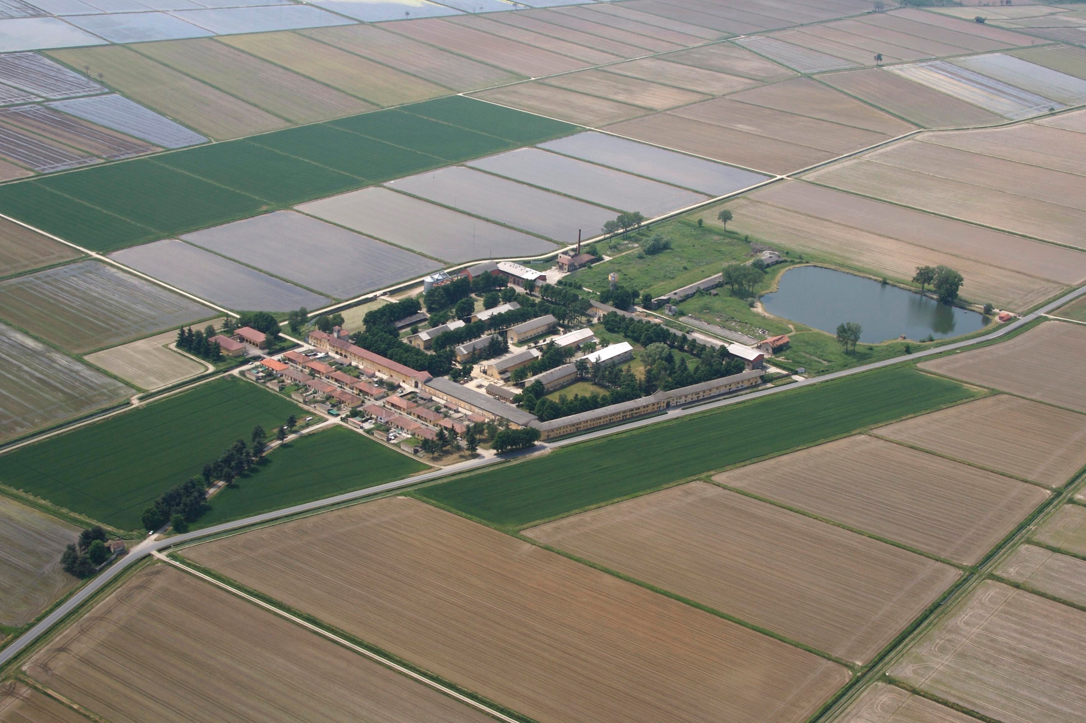 Luftaufnahme von Reisfeldern, teils geflutet, und einem grossen Hofkomplex mit Gebäuden und einem See.
