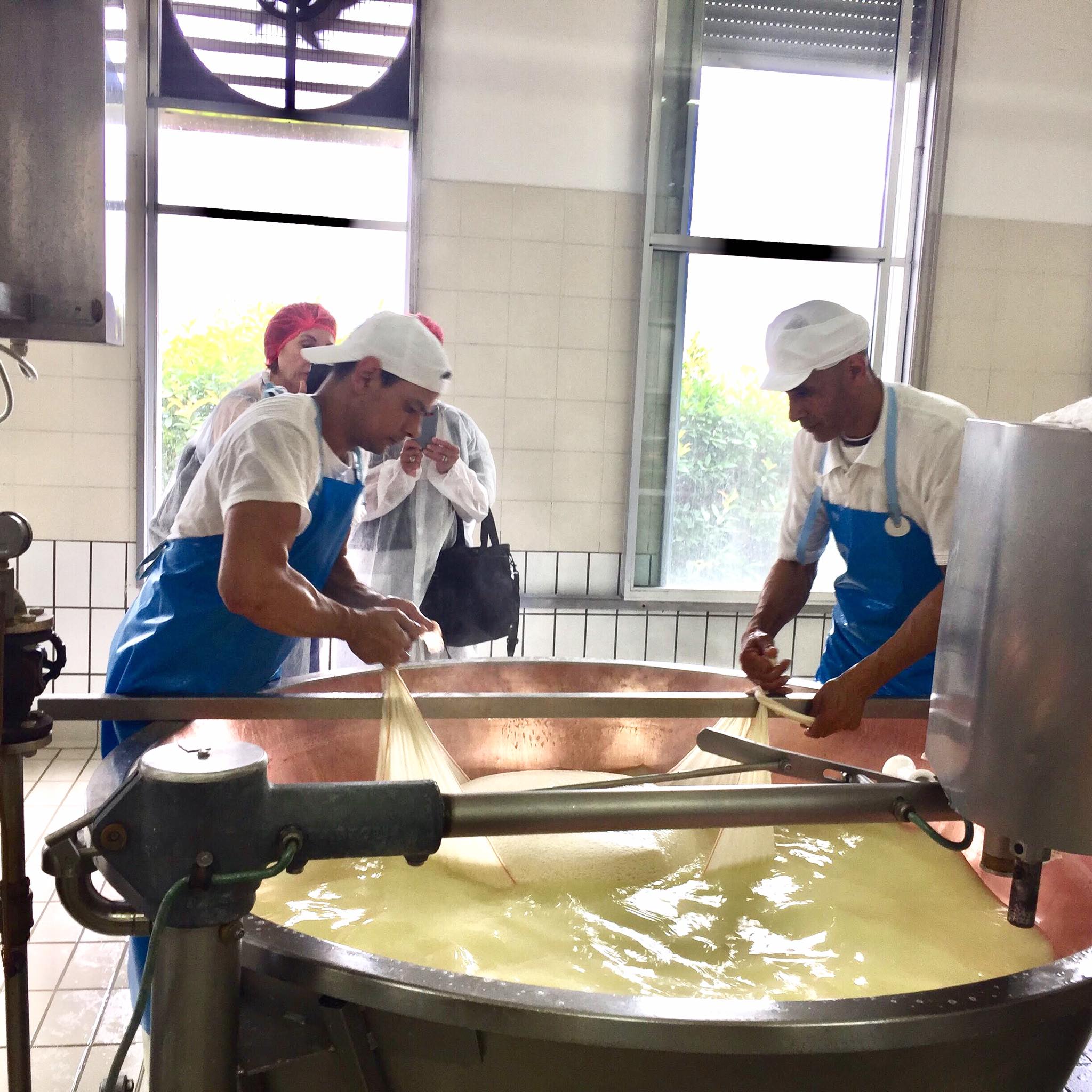 Zwei Käser in blauen Schürzen und weissen Kappen arbeiten an einem grossen Kupferkessel mit Käsebruch und Molke.