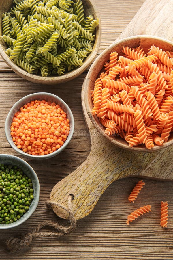 Grüne und orange Fusilli-Pasta in Holzschalen, dazu rote Linsen und Mungbohnen in Keramikschalen auf rustikalem…