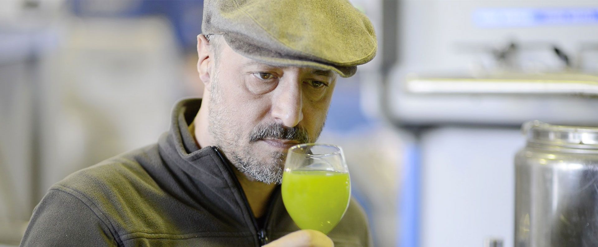 Mann mit Bart und Mütze riecht konzentriert an einem Glas mit grünem Olivenöl in einer Produktionshalle.