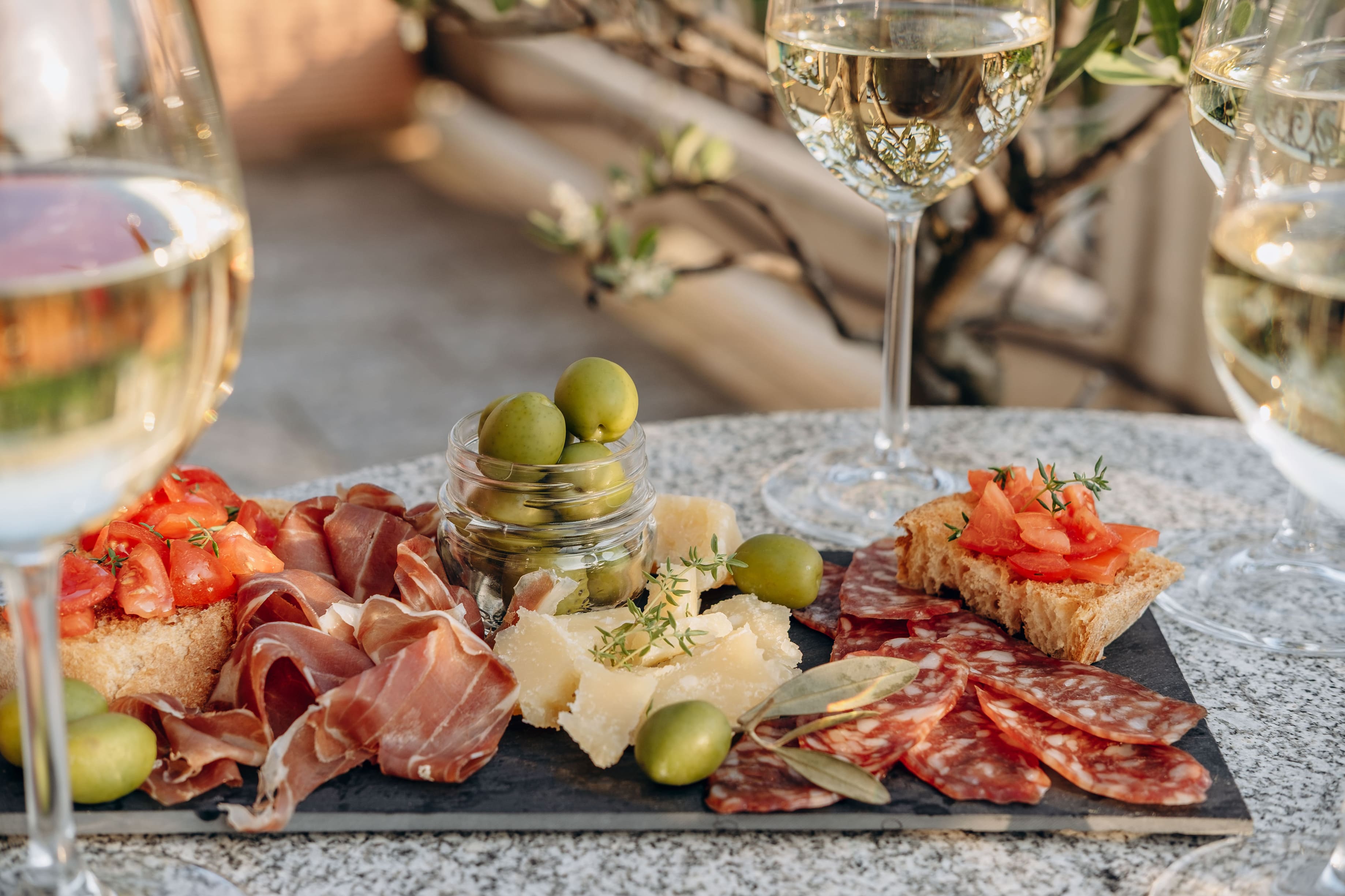 Schieferplatte mit Prosciutto, Salami, Käse, Oliven und Tomaten-Bruschetta. Weissweingläser auf Granittisch im Freien.