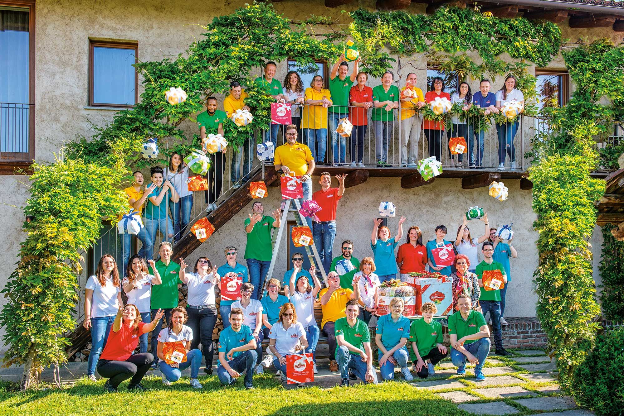 Zahlreiche Mitarbeitende in farbigen Shirts vor einem Gebäude mit Balkon. Sie halten und werfen festliche…