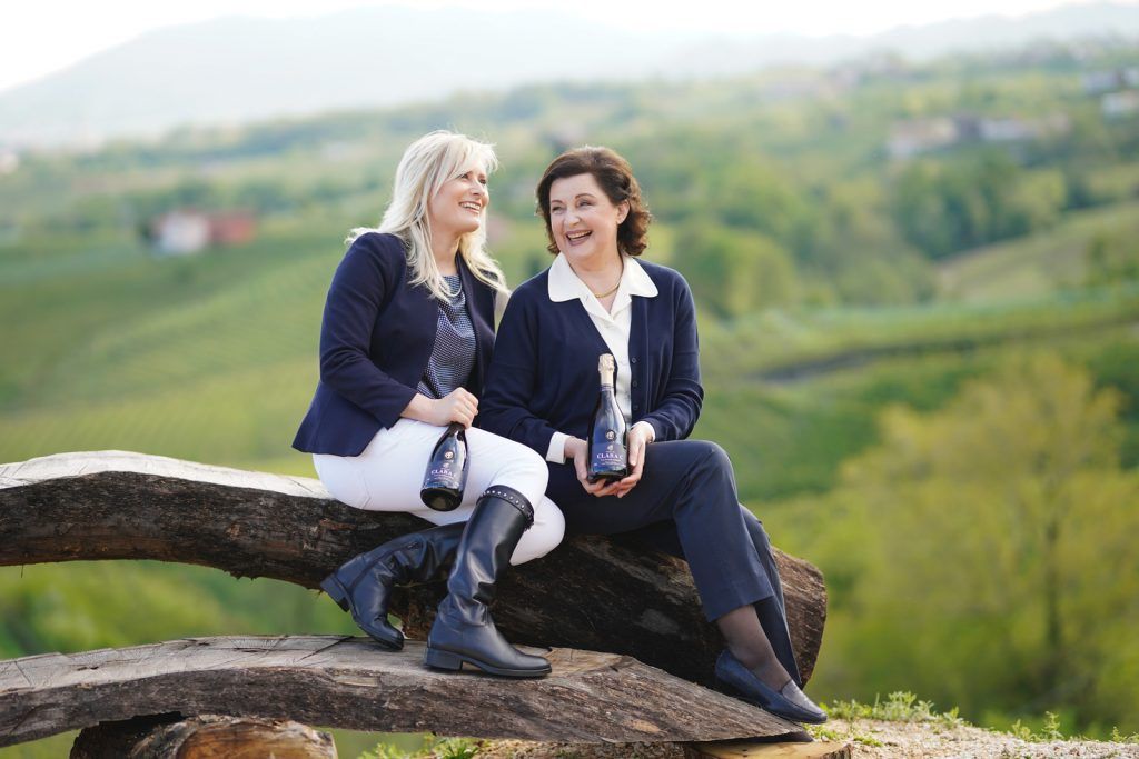 Zwei Frauen sitzen lächelnd auf einem Holzstamm, halten Flaschen von Clara C Sekt in einer grünen Weinberglandschaft.