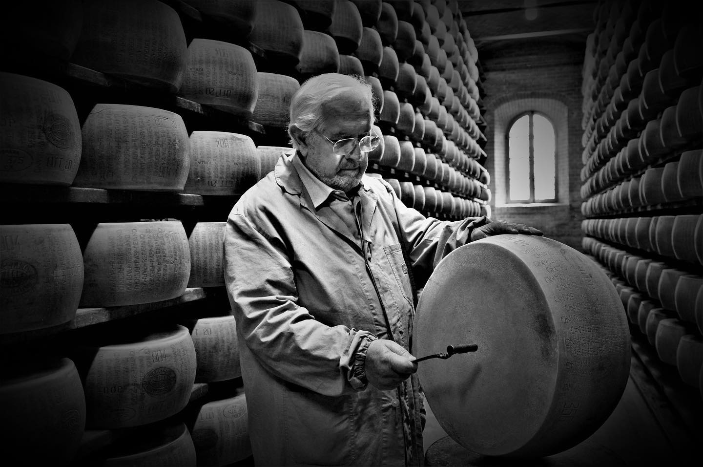 Mann mit Brille und Arbeitskittel prüft einen Parmigiano Reggiano Käselaib in einem Reifekeller voller Käse.