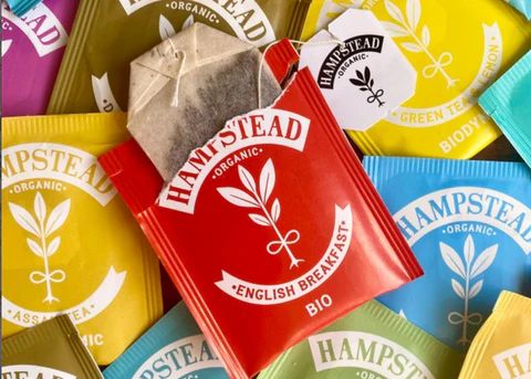 Verschiedene Hampstead Organic Teebeutel und Packungen in Rot, Gelb, Blau, Grün und Violett. Ein offener Beutel zeigt…